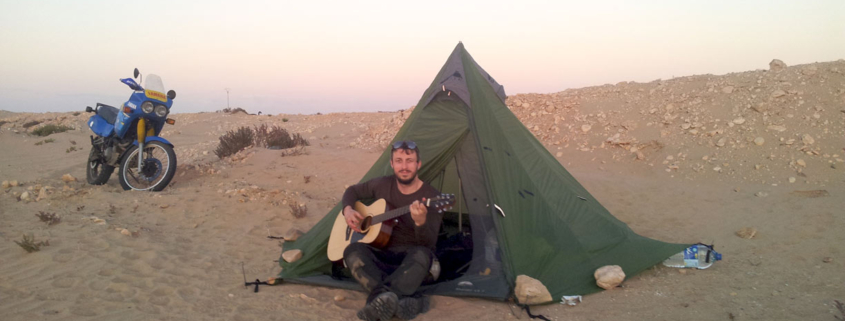 Man playing a Voyage Air guitar outside a tent in a desert campsite with a motorcycle nearby during sunset.