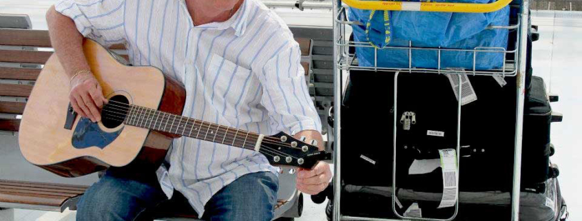 Man playing a Voyage Air guitar while sitting at an airport with luggage, demonstrating the convenience of a travel-friendly folding guitar.