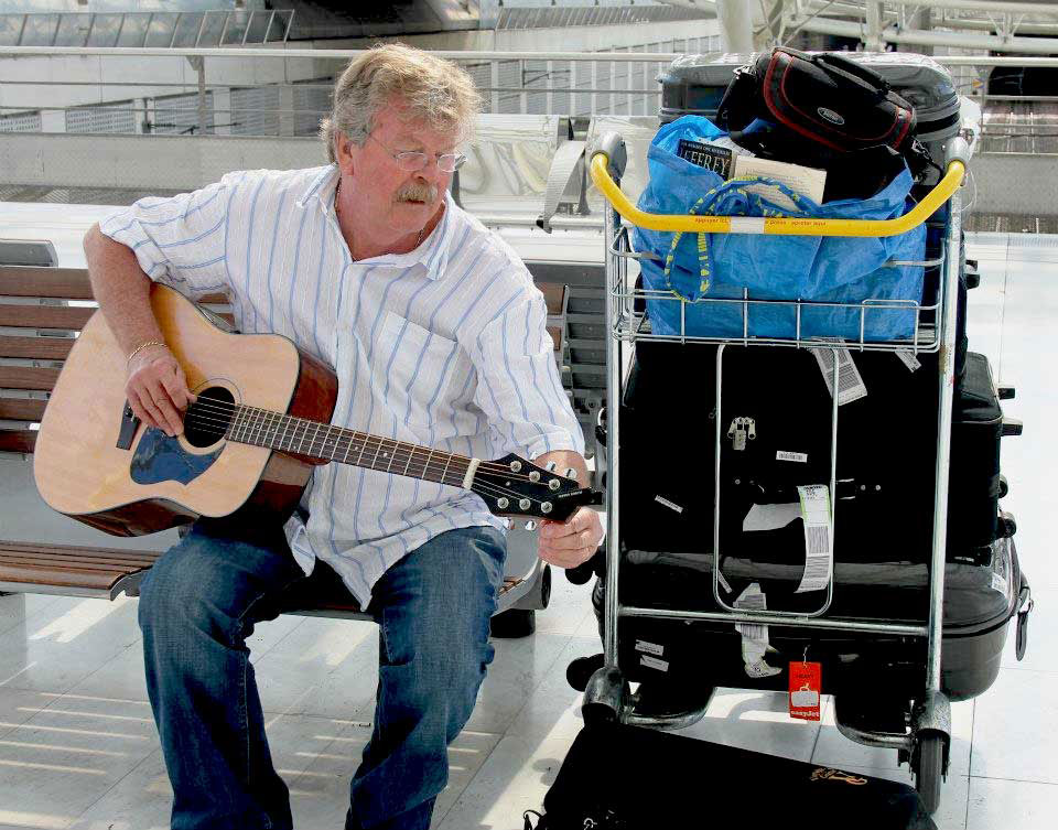 Man playing a Voyage Air guitar while sitting at an airport with luggage, demonstrating the convenience of a travel-friendly folding guitar.