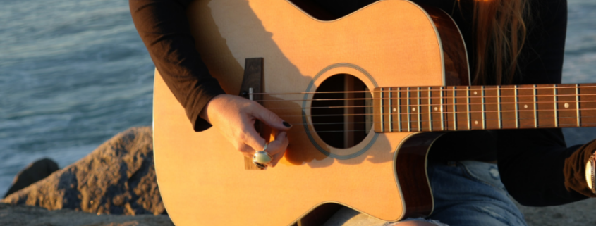 Person playing an acoustic guitar outdoors near the water, highlighting proper handling and care to keep a travel-friendly instrument in great condition.