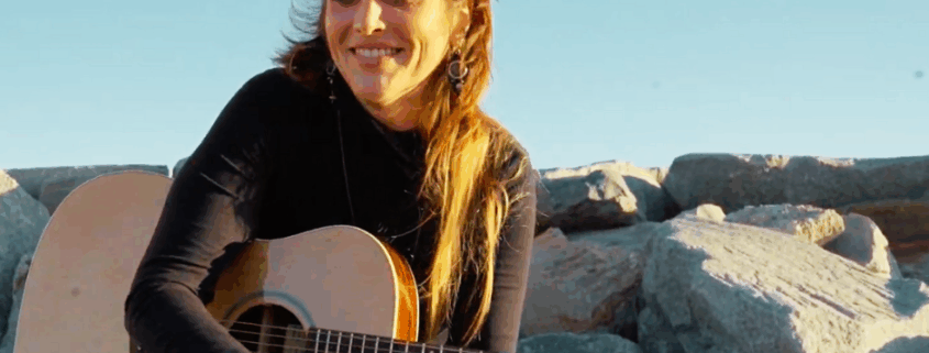 A woman playing a Voyage Air guitar while sitting on rocks outdoors, smiling in natural sunlight.